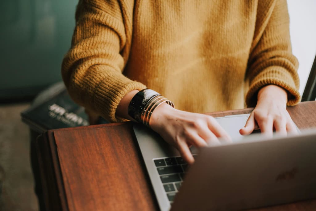 Person in a mustard sweater and gold bracelets typing on a laptop at a wooden table