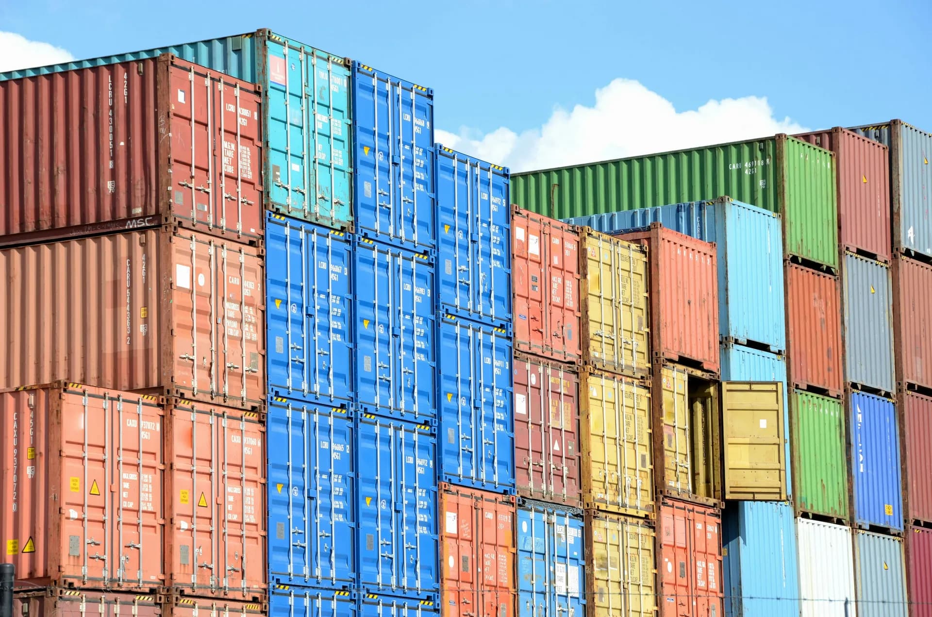 Stacks of red, blue, yellow, and green shipping containers at a port under a blue sky with clouds