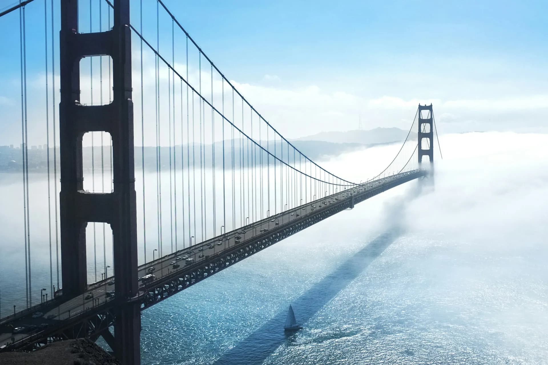 Golden Gate Bridge stretching across the bay with traffic, its far tower rising out of low fog above a small sailboat